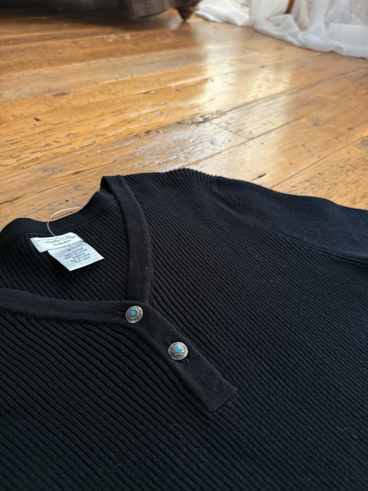 The Walhalla Black Ribbed Dress by Savannah Sevens western life{&}style, featuring long sleeves, a V-neck, and two decorative buttons, is laid flat on a wooden floor with white curtains and sunlight in the background.