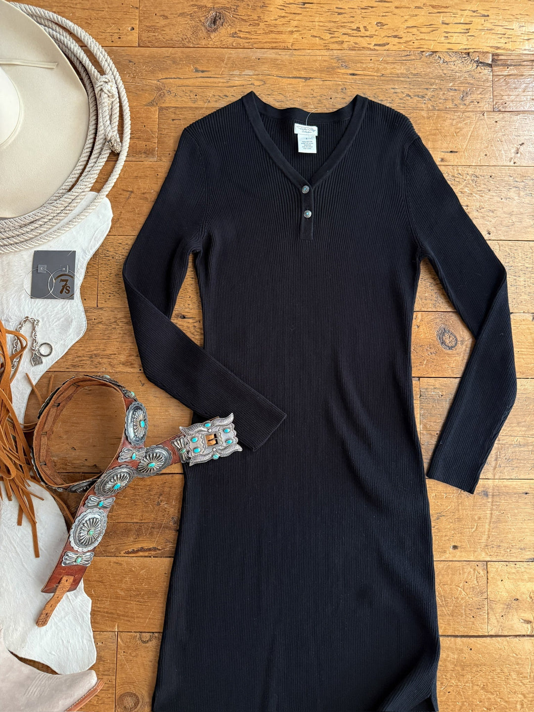 The Walhalla Black Ribbed Dress by Savannah Sevens western life{&}style is laid flat on a wooden floor, paired with a tan hat, fringed brown bag, multicolored embroidered belt, and jewelry for a versatile, stylish look.