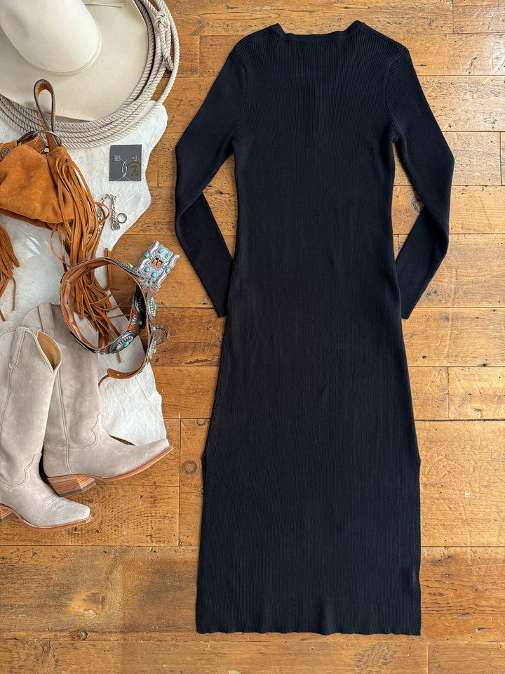 The Walhalla Black Ribbed Dress by Savannah Sevens western life{&}style is laid flat on a wooden floor, styled with beige cowboy boots, a tan hat, suede fringe bag, belts, jewelry, and a wallet for a complete western look.