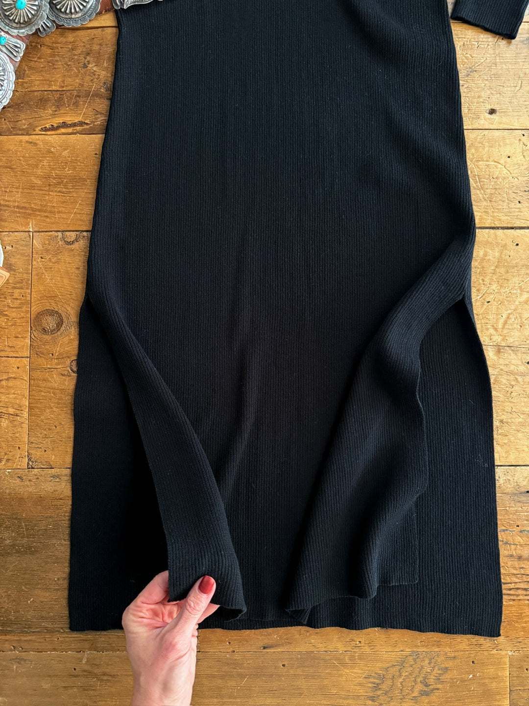A hand lifts the hem of the Savannah Sevens western life{&}style Walhalla Black Ribbed Dress laid flat on wood. A silver concho belt appears in the top left, accentuating the classic look of this long sleeve dress.