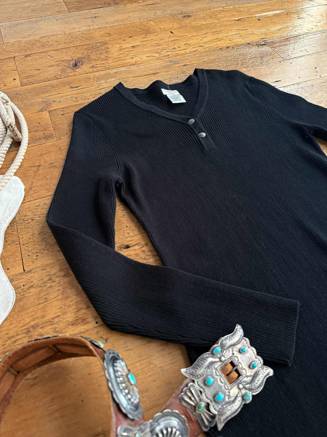 The Walhalla Black Ribbed Dress by Savannah Sevens western life{&}style is laid out on a wooden floor beside a tooled leather belt with a large silver buckle and a coiled rope.