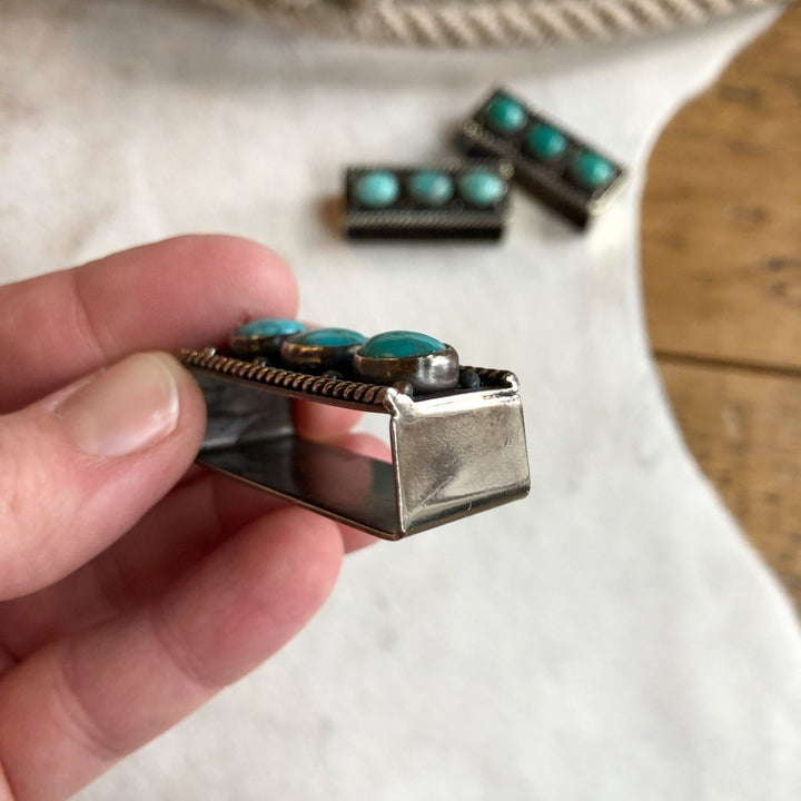 A hand holds the Wallowa Belt Keeper by Savannah Sevens western life{&}style; two more turquoise-accented pieces from the brand are displayed on a soft white surface in the background.