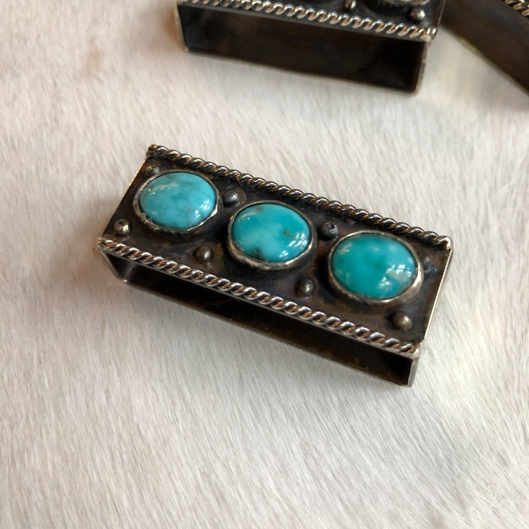 The Wallowa Belt Keeper by Savannah Sevens western life{&}style features a rectangular German silver design with three round turquoise stones, twisted wire accents, and metallic beads, displayed on a white textured surface.
