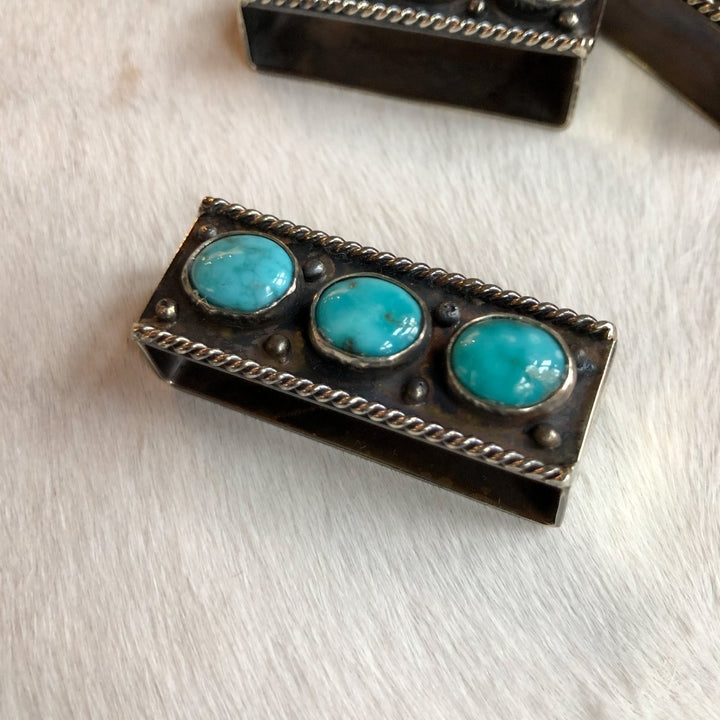 The Wallowa Belt Keeper by Savannah Sevens western life{&}style features a rectangular German silver design with three round turquoise stones, twisted wire accents, and metallic beads, displayed on a white textured surface.