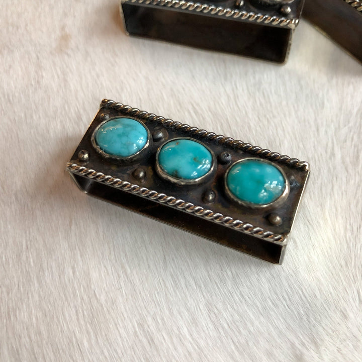 The Wallowa Belt Keeper by Savannah Sevens western life{&}style features a rectangular German silver design with three round turquoise stones, twisted wire accents, and metallic beads, displayed on a white textured surface.