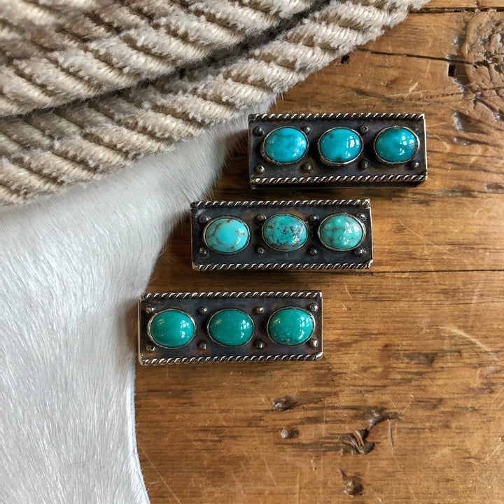 Three Wallowa Belt Keepers by Savannah Sevens western life{&}style are displayed on a wooden surface, accompanied by a striped fabric and a cowhide.