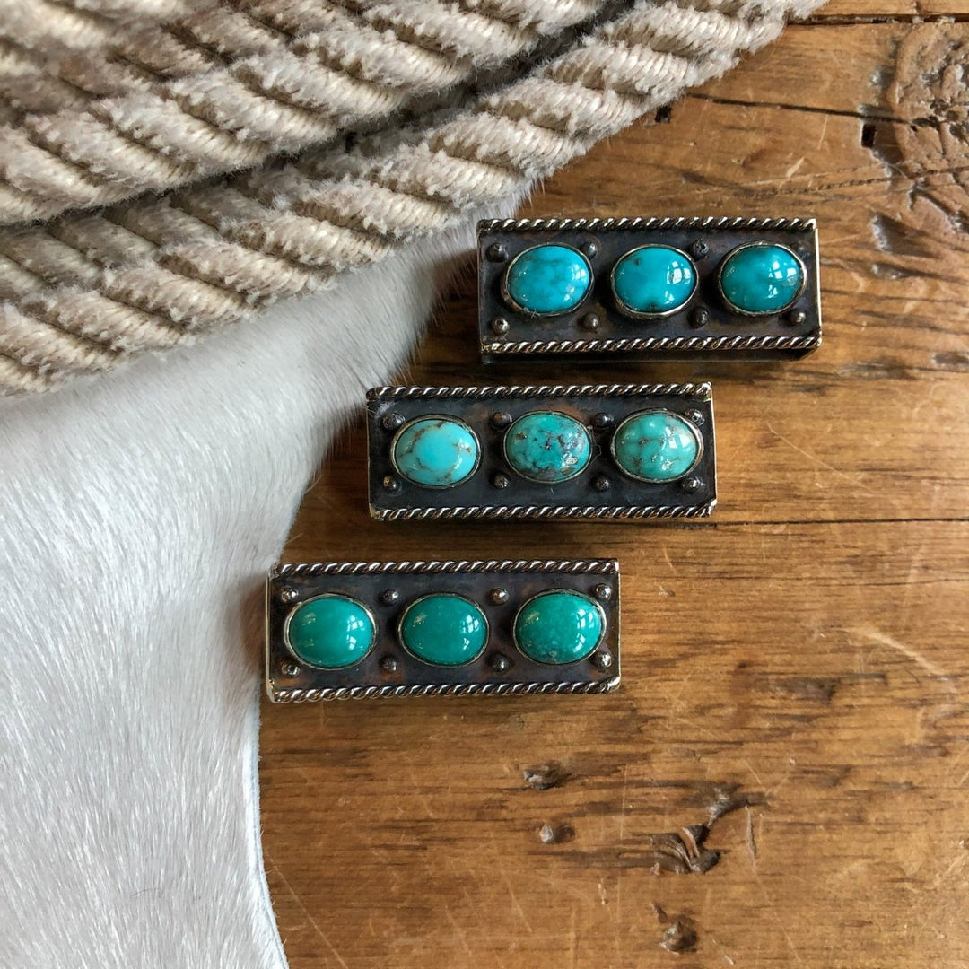 Three Wallowa Belt Keepers by Savannah Sevens western life{&}style are displayed on a wooden surface, accompanied by a striped fabric and a cowhide.
