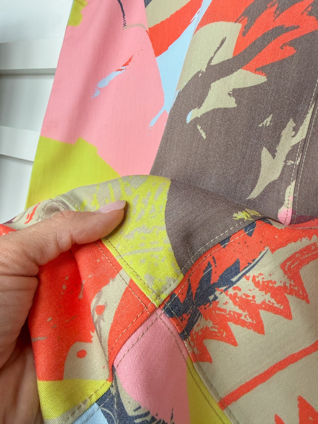 A hand holds the Wanette Abstract Boot Graphic Dress by Savannah Sevens western life{&}style, an oversized short sleeve dress featuring a colorful design with pink, yellow, orange, gray, and blue abstract shapes and visible seam stitching.