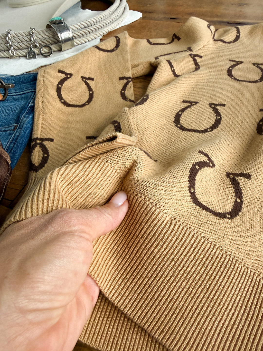 A hand holds the beige Wapato Horseshoe Cardigan from Savannah Sevens western life&style, featuring brown horseshoe knit patterns. Next to it on a wooden surface are blue jeans, a rope belt, and a white hat.