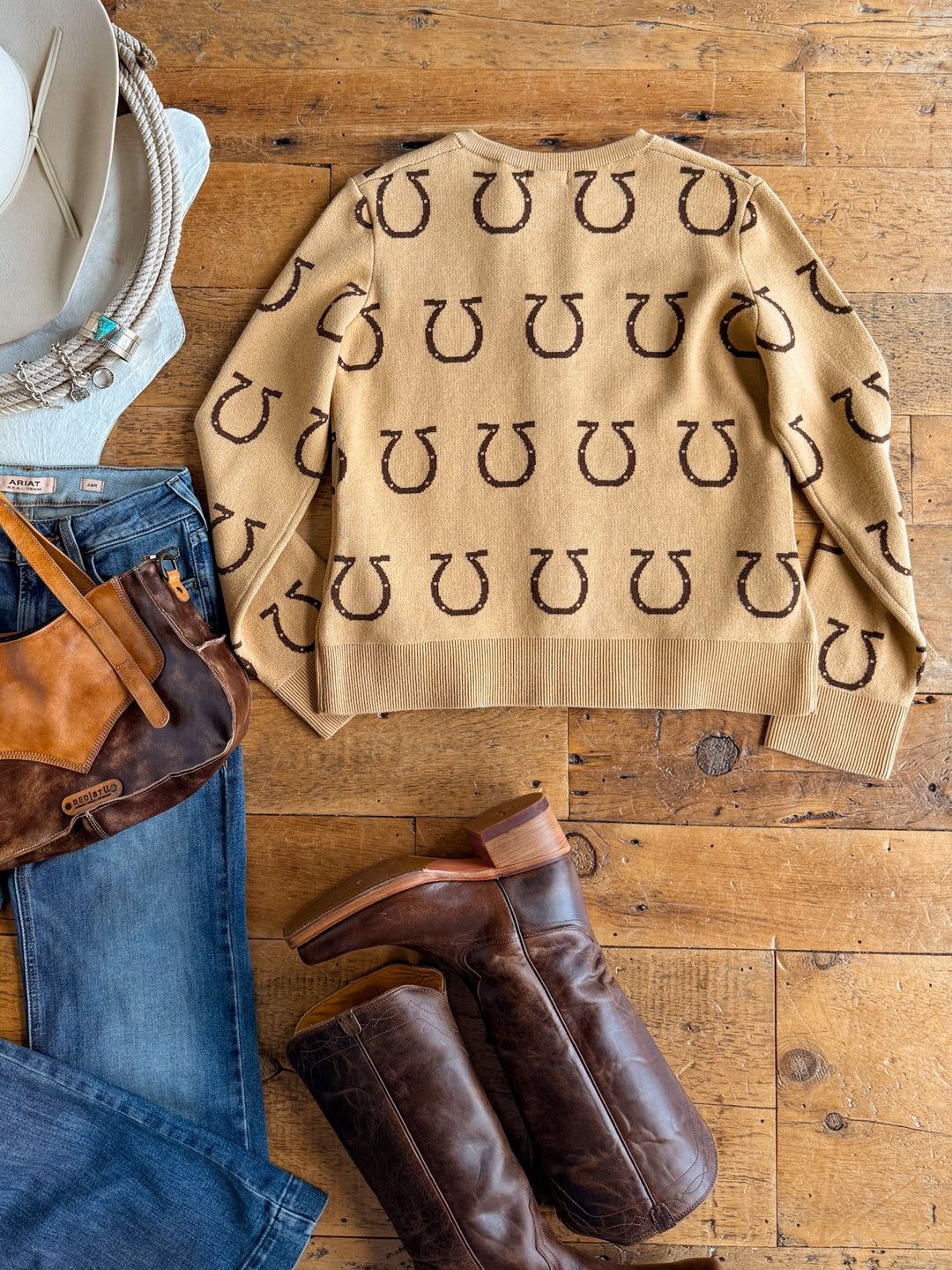 Flat lay of a tan Savannah Sevens western life{&}style Wapato Horseshoe Cardigan with brown horseshoe knit prints, paired with blue jeans, brown leather boots, a brown suede bag, a white hat, and turquoise jewelry on a wooden floor.