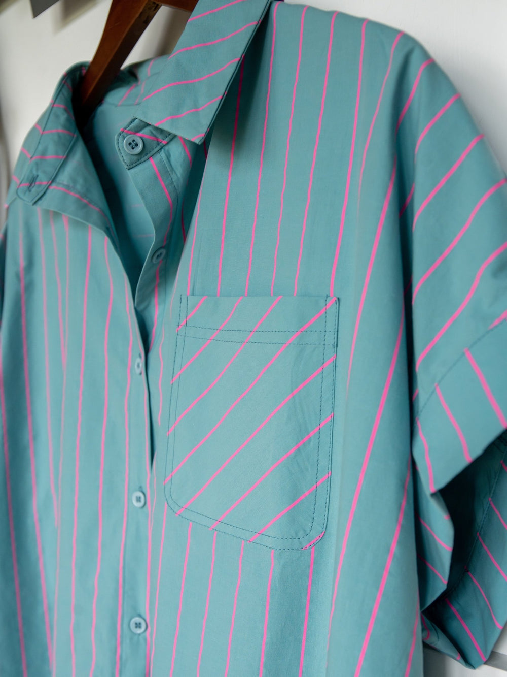The Wapello Turquoise Blue Button Up Top by Savannah Sevens western life{&}style features short sleeves, pink diagonal stripes, a front pocket, and an oversized fit for comfort and style. Shown hanging on a wooden hanger against a white background.