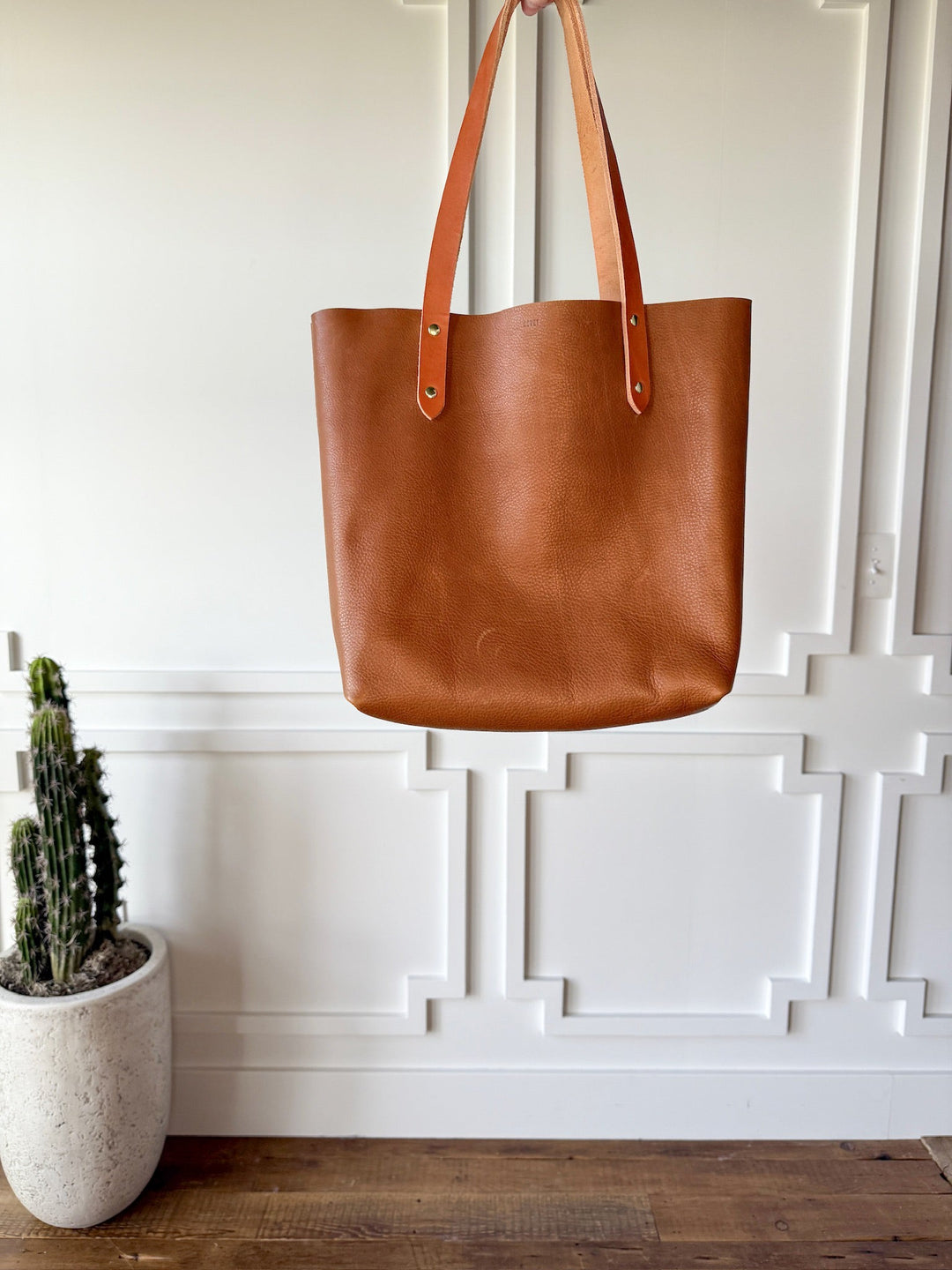 The Wasco by Savannah Sevens western life{&}style, a full-grain pecan leather tote with two handles, hangs before a white paneled wall while a potted cactus rests on the wooden floor nearby, adding a touch of elegance to the space.
