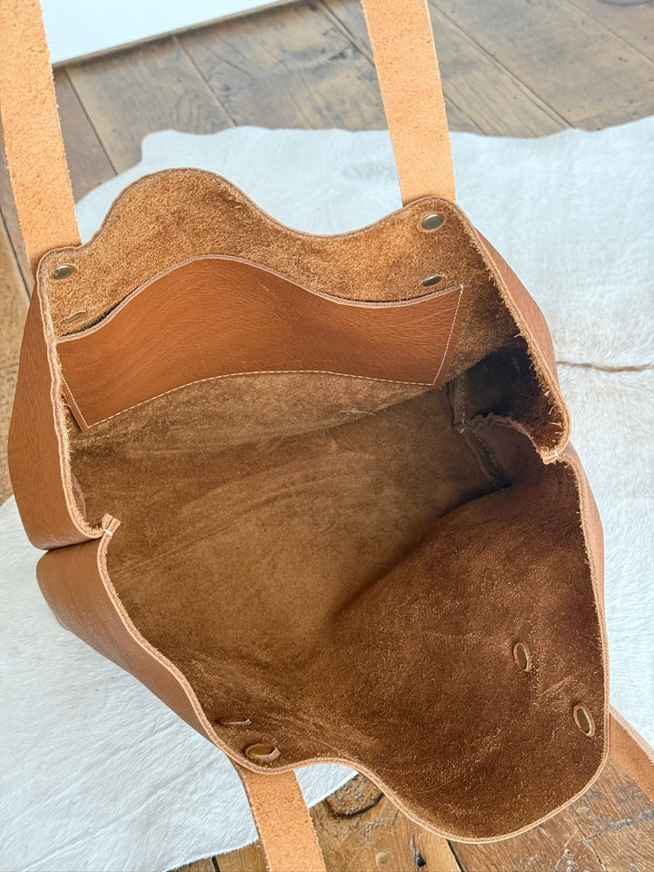 The Wasco tote by Savannah Sevens western life{&}style features open pecan full-grain leather, a suede interior, and a small inner pocket. Made in California, it rests on wood and white fabric with its spacious compartment partially revealed. By Covet. 
