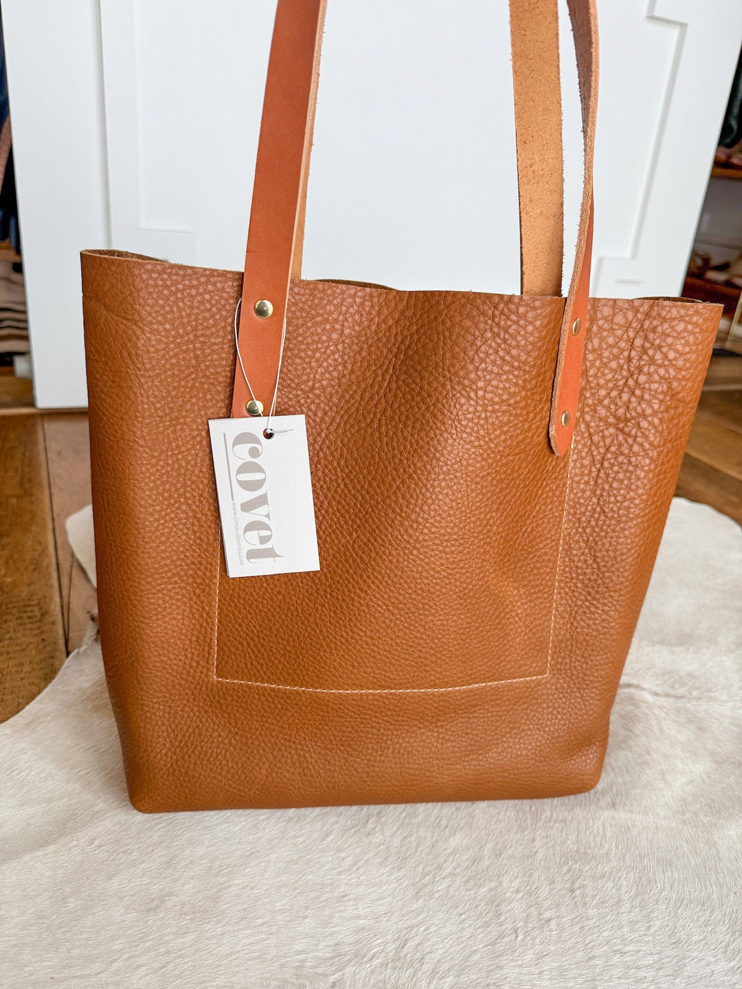 The Wasco by Savannah Sevens western life{&}style is a full-grain pecan leather tote with sturdy handles, a front pocket, and a brass-riveted "covet" tag—shown on a light rug. Crafted for lasting quality. By Covet. 