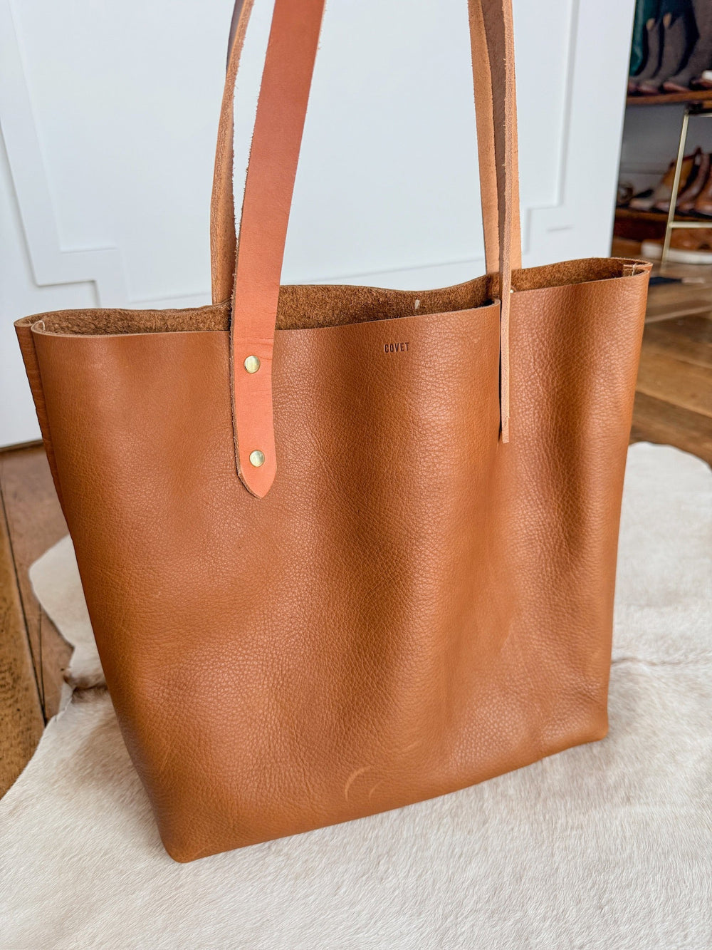 The Wasco by Savannah Sevens western life{&}style, a pecan leather tote with two flat handles made in California, rests on a light cowhide rug in a bright room with wood floors and white walls. By Covet. 