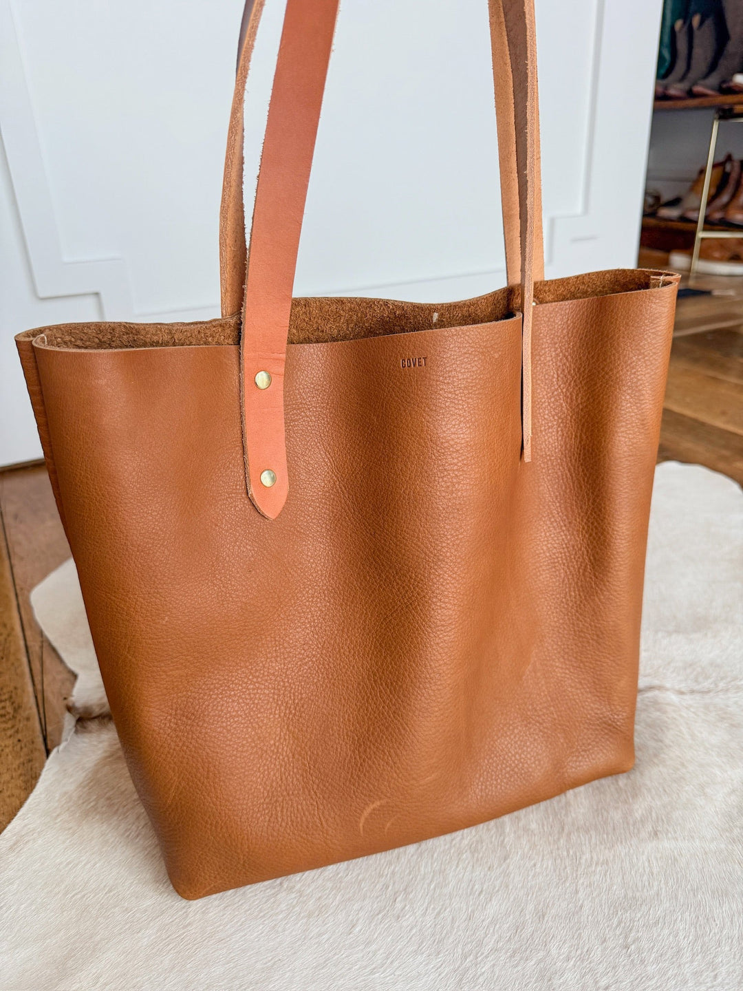 The Wasco by Savannah Sevens western life{&}style, a pecan leather tote with two flat handles made in California, rests on a light cowhide rug in a bright room with wood floors and white walls. By Covet. 