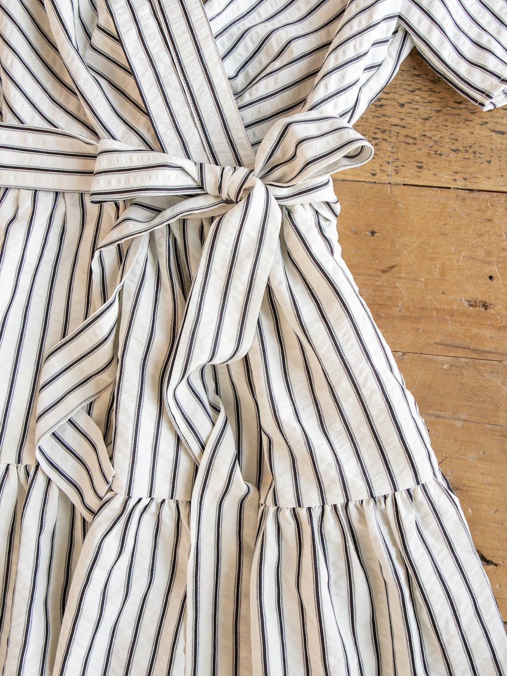 The Wasilla Off-White Striped Textured Dress by Savannah Sevens western life{&}style, crafted from a cotton blend with black vertical stripes on white fabric, features short puff sleeves, a belted waist tied in a knot, and a ruffled skirt. It is displayed laid out on a wooden surface.
