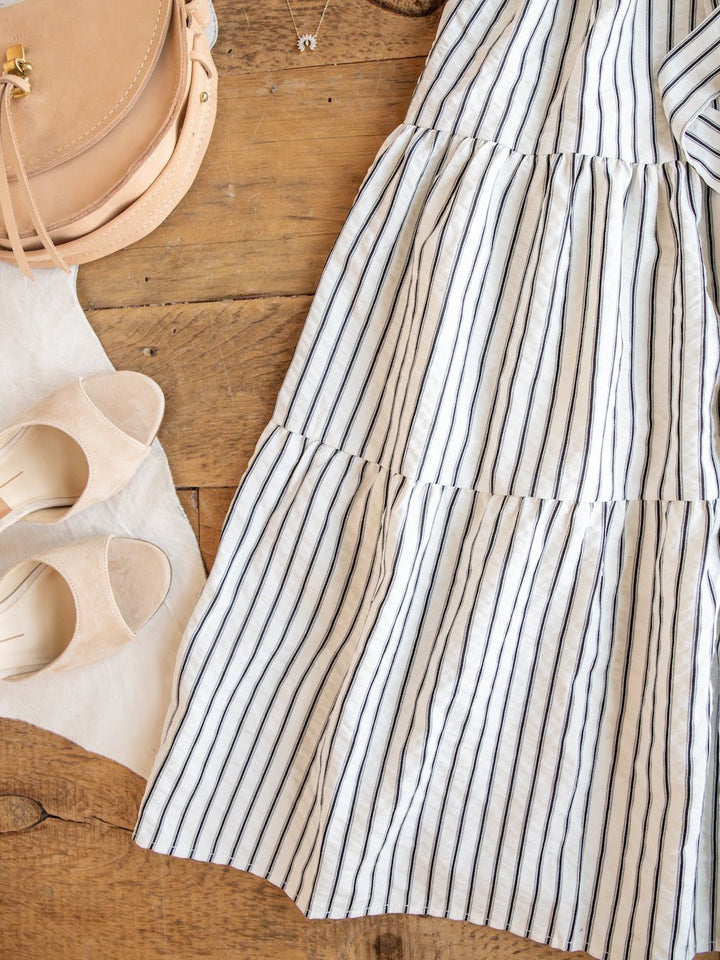 The Wasilla Off-White Striped Textured Dress by Savannah Sevens western life{&}style, paired with beige open-toe sandals and a tan leather purse, are arranged on a rustic wooden floor.