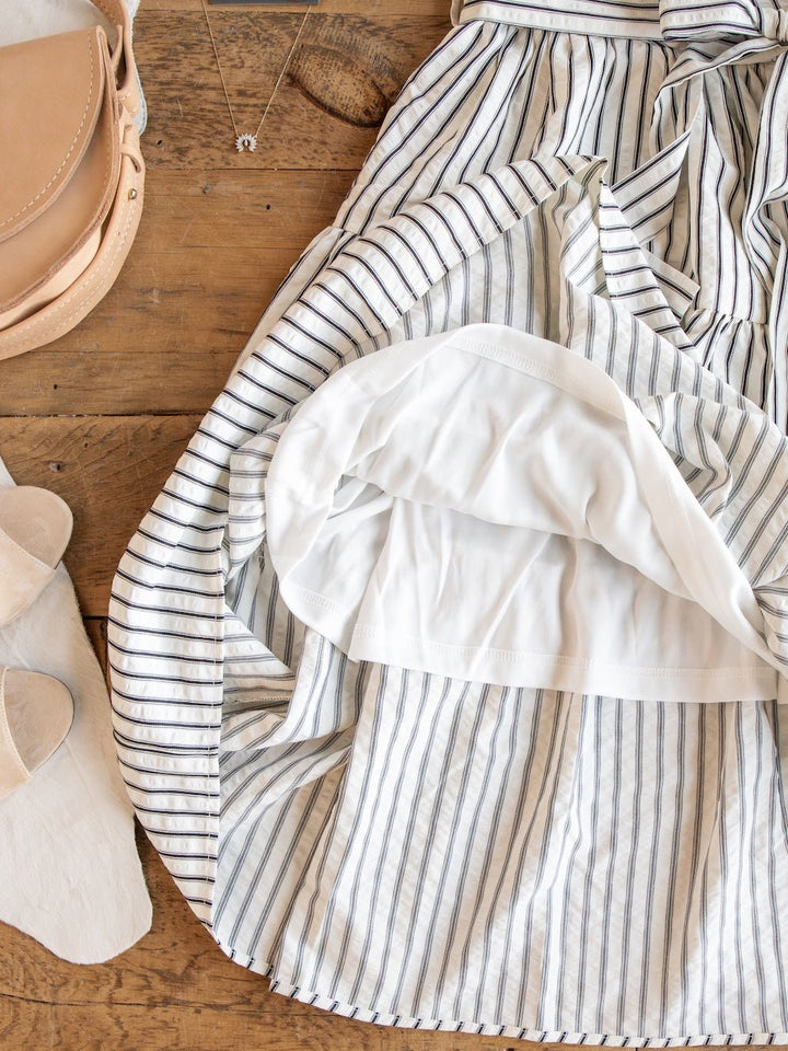 The Wasilla Off-White Striped Textured Dress by Savannah Sevens western life{&}style is displayed on a wooden surface with its white lining visible, styled alongside a tan bag, a necklace, and light-colored slide sandals for an effortless, relaxed look.