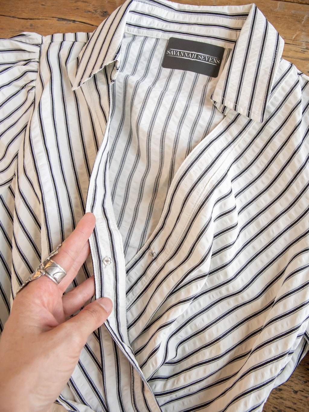 A hand adorned with silver rings holds the edge of the Wasilla Off-White Striped Textured Dress, which features black vertical stripes and a label inside the collar that reads Savannah Sevens western life{&}style. The dress is laid flat on a wooden surface.