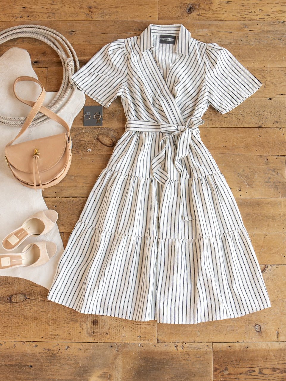The Wasilla Off-White Striped Textured Dress by Savannah Sevens western life{&}style is displayed on a wooden floor, styled with a beige purse, a pair of sandals, a tote bag, and coordinating earrings arranged neatly beside it.