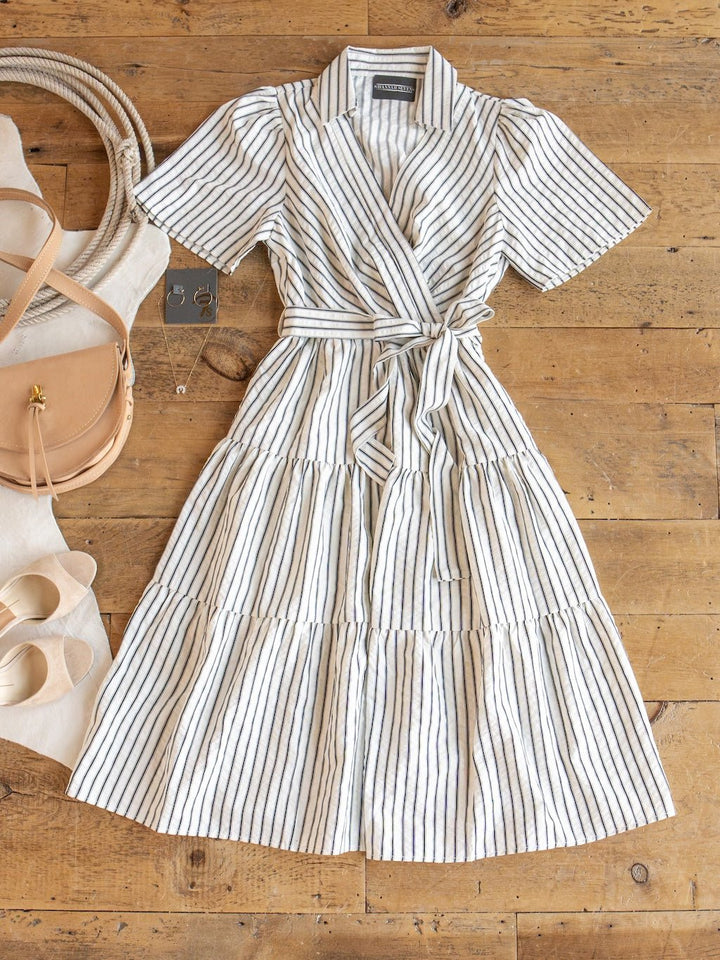 The Wasilla Off-White Striped Textured Dress by Savannah Sevens western life{&}style, a short-sleeved cotton blend wrap dress with thin black stripes, is displayed on a wooden floor alongside a tan purse, cream heels, a white bag, and a pair of earrings.