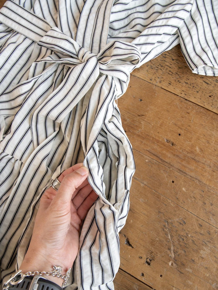 A hand reveals the hidden pocket of the Wasilla Off-White Striped Textured Dress by Savannah Sevens western life{&}style, featuring a cotton-blend fabric and waist tie, displayed on a wooden surface.