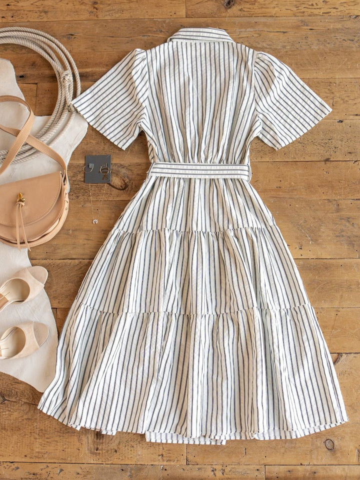 The Wasilla Off-White Striped Textured Dress by Savannah Sevens western life{&}style—a white, short-sleeve, collared cotton-blend midi dress with black vertical stripes—is laid out on a wooden floor, surrounded by beige shoes, a tan purse, a white bag, and black earrings.