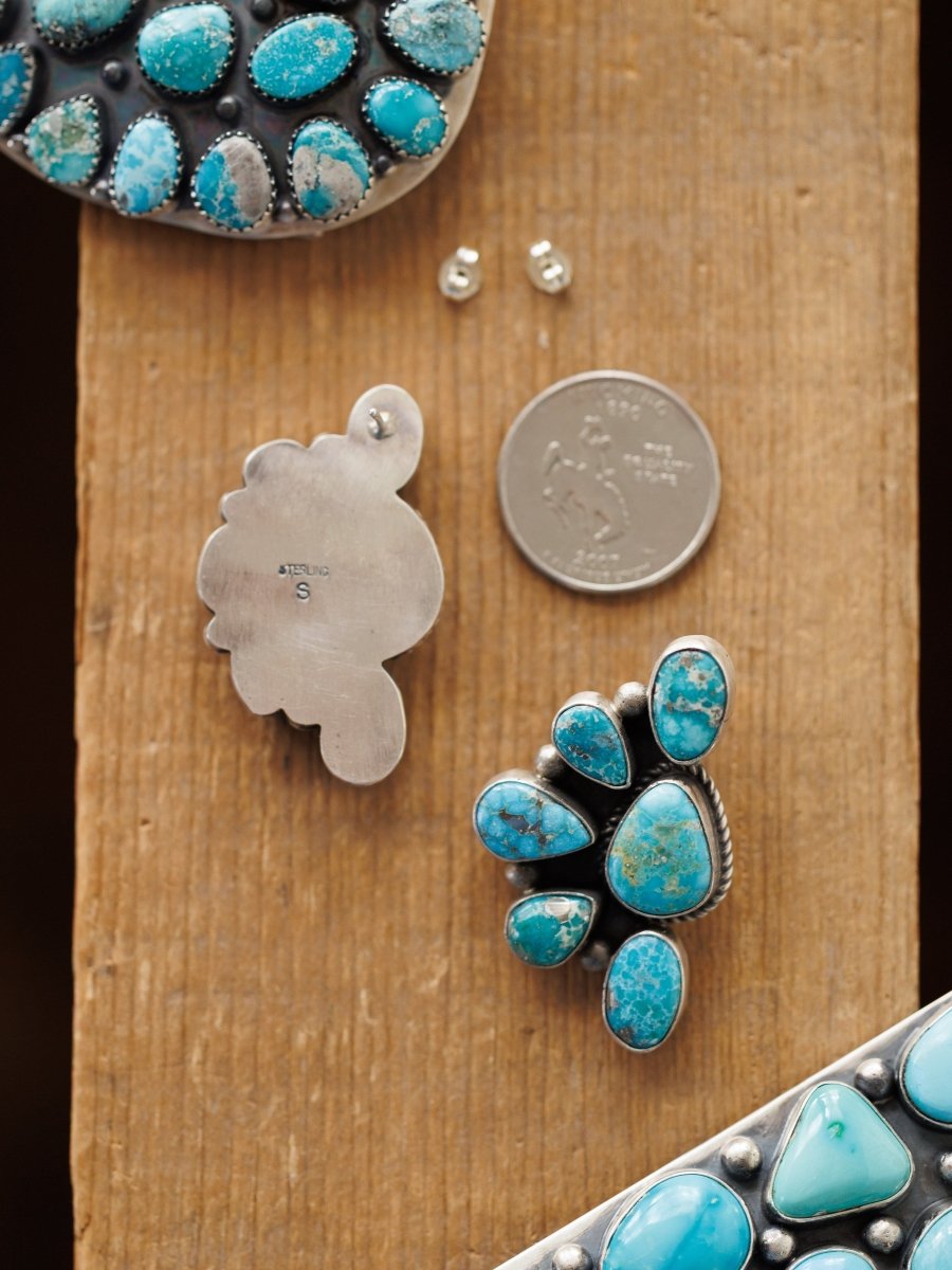 The Watseka Earrings by Savannah Sevens western life{&}style, along with a silver pendant, earring backs, and a U.S. quarter, rest on a wooden surface. The Native American handcrafted turquoise pieces feature unique cluster designs set in silver.