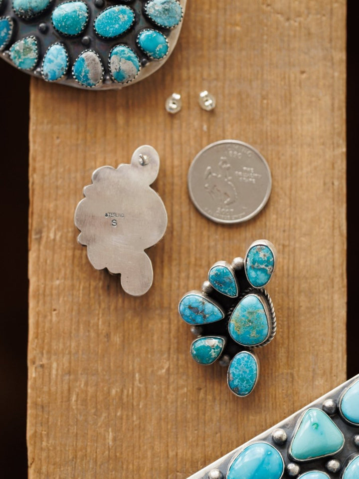 The Watseka Earrings by Savannah Sevens western life{&}style, along with a silver pendant, earring backs, and a U.S. quarter, rest on a wooden surface. The Native American handcrafted turquoise pieces feature unique cluster designs set in silver.