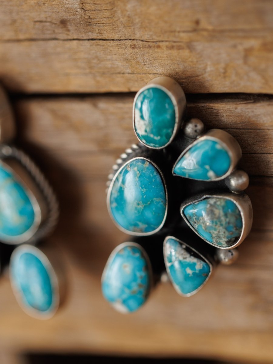 A close-up of the Watseka Earrings by Savannah Sevens western life{&}style, featuring five irregularly shaped turquoise stones and handcrafted in a style reminiscent of Native American artistry, resting on a rustic wooden surface.