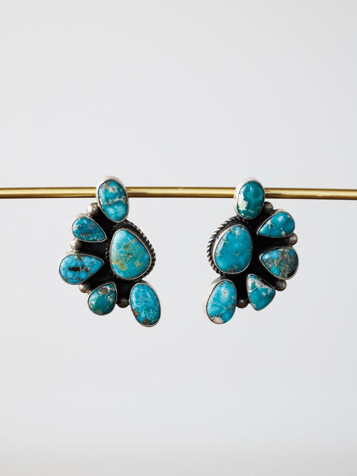 The Watseka Earrings by Savannah Sevens western life{&}style are Native American handcrafted sterling silver earrings featuring turquoise stones in a fan shape, displayed hanging from a gold rod on a plain white background.