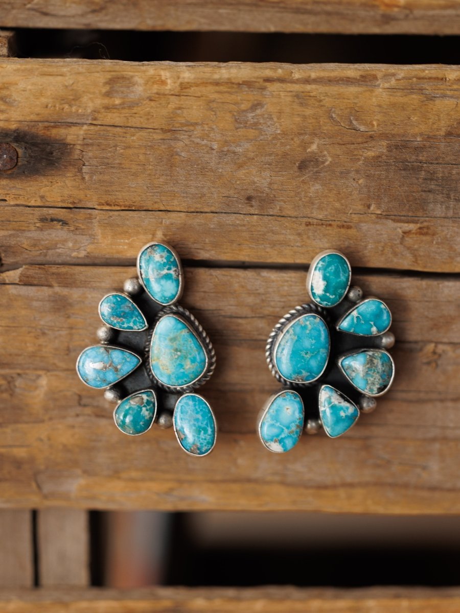 The Watseka Earrings by Savannah Sevens western life{&}style are handcrafted Native American sterling silver earrings with multiple turquoise stones in a circular floral design, shown on a rustic wooden background.