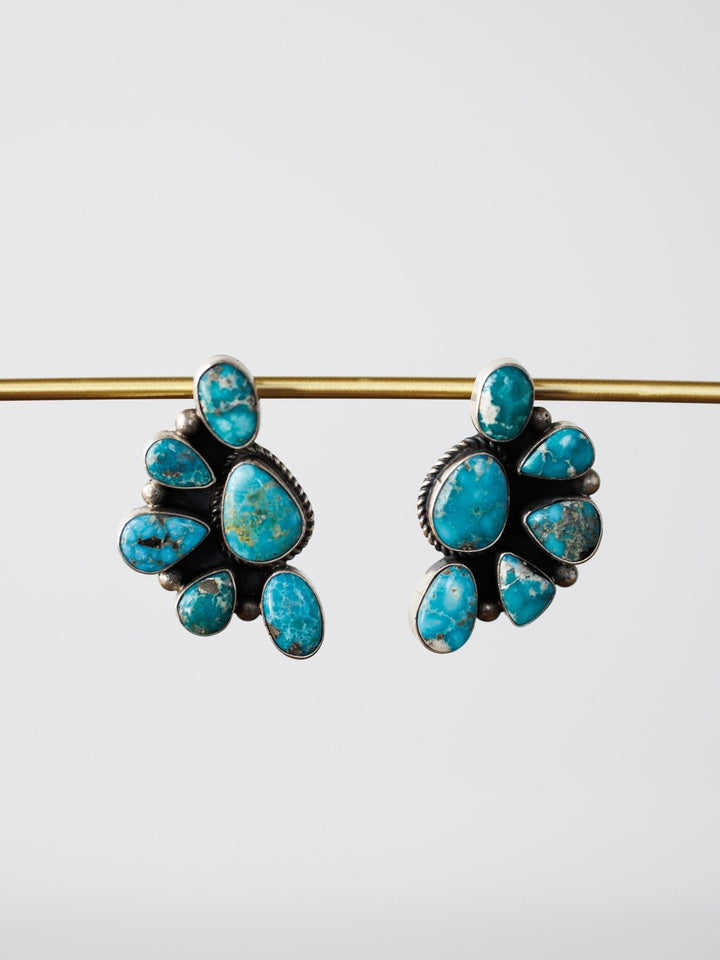 The Watseka Earrings by Savannah Sevens western life{&}style are Native American handcrafted sterling silver earrings featuring turquoise stones in a fan shape, displayed hanging from a gold rod on a plain white background.
