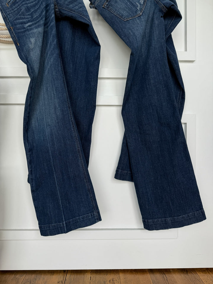 Two pairs of Savannah Sevens western life{&}style The Waylynn Perfect Rise Trouser Jean *Exclusive* in medium dark wash hang side by side over a white door, legs extending downward and the wooden floor partially visible below.