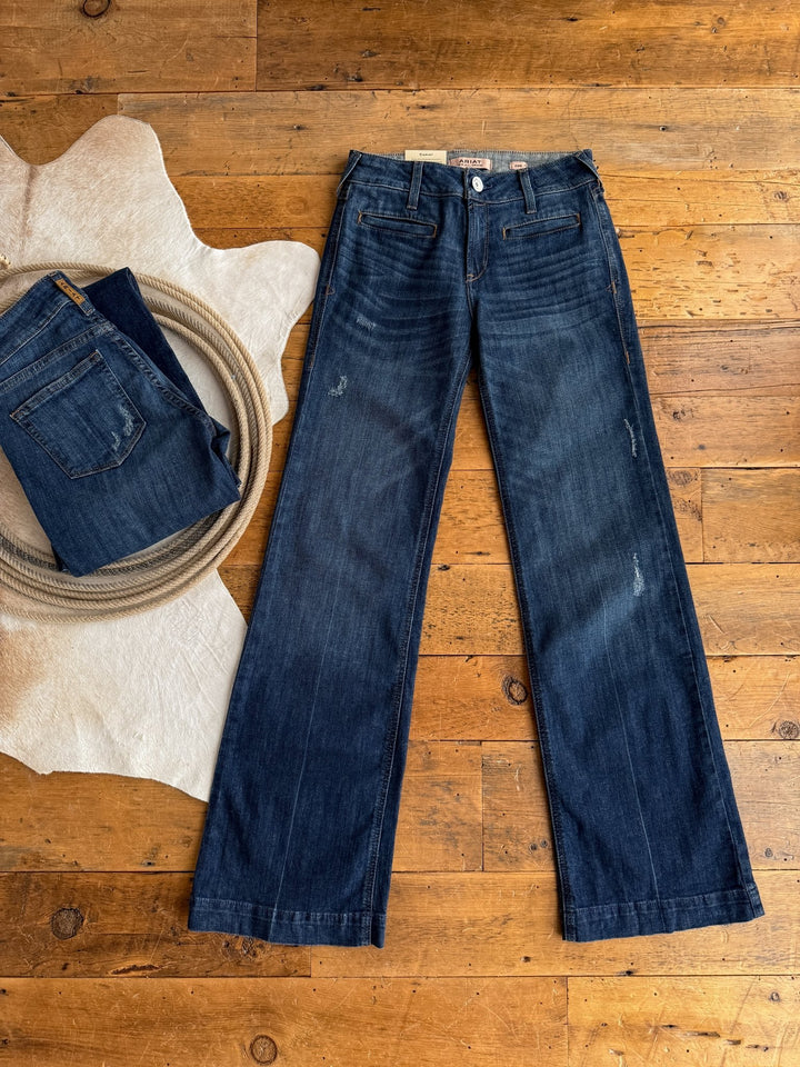 Two pairs of Savannah Sevens The Waylynn Perfect Rise Trouser Jeans are shown on a wooden floor: one is laid flat, while the other, with a no-gap waist, is folded and placed on a light-colored rug beside a coiled rope.