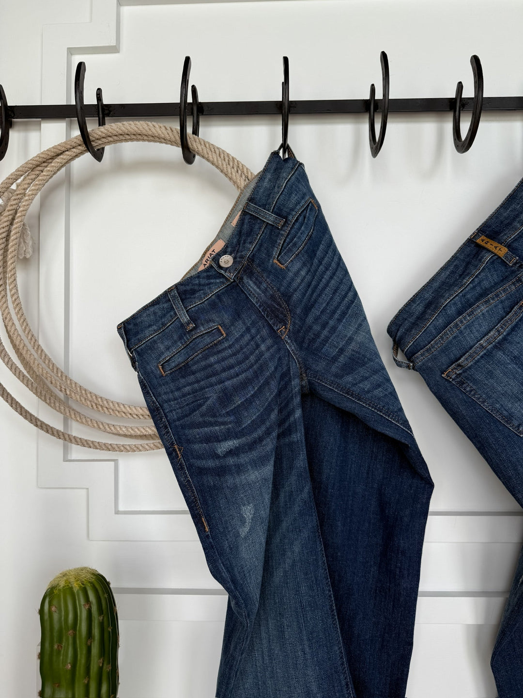 The Waylynn Perfect Rise Trouser Jean by Savannah Sevens western life{&}style hangs on a black hook rack next to another pair of jeans, a looped rope, and a green cactus beneath on a white paneled wall with no-gap waist styling.