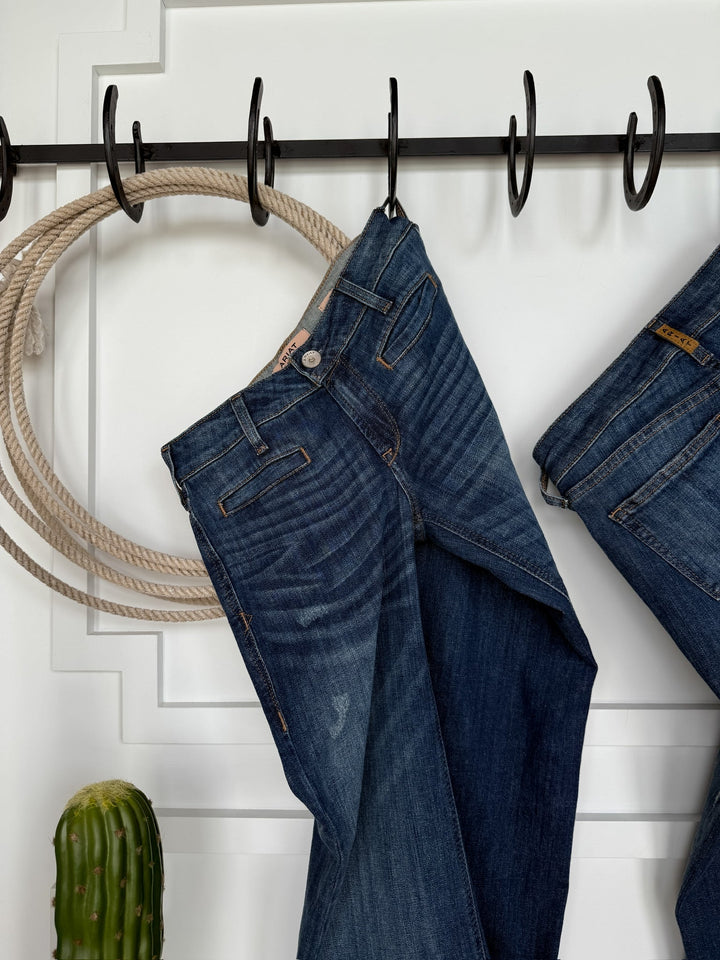 The Waylynn Perfect Rise Trouser Jean by Savannah Sevens western life{&}style hangs on a black hook rack next to another pair of jeans, a looped rope, and a green cactus beneath on a white paneled wall with no-gap waist styling.
