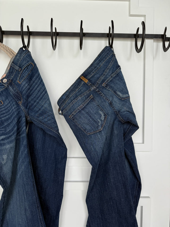 Two pairs of The Waylynn Perfect Rise Trouser Jeans by Savannah Sevens western life{&}style hang by their belt loops on black metal hooks against a white paneled wall, showcasing no-gap waistbands, back pockets, and detailed seams.
