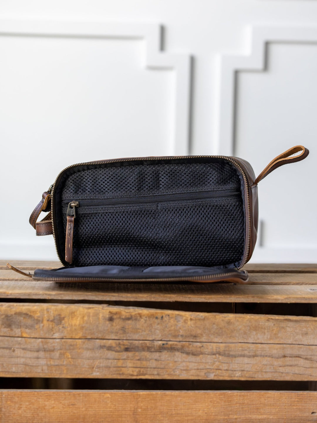 The Wayneboro Leather Shave Bag by Savannah Sevens western life{&}style, a brown leather dopp kit with a zipper, is open to reveal a black mesh interior pocket. It rests on wood with a white paneled wall in the background.