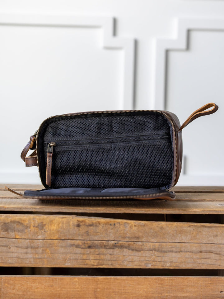 The Wayneboro Leather Shave Bag by Savannah Sevens western life{&}style, a brown leather dopp kit with a zipper, is open to reveal a black mesh interior pocket. It rests on wood with a white paneled wall in the background.