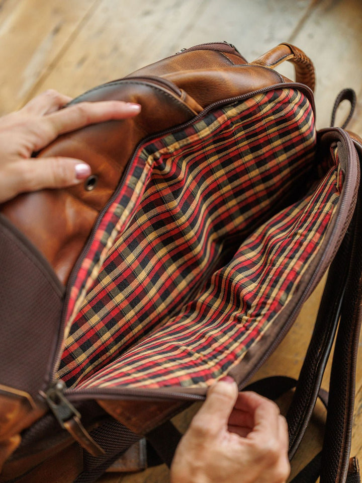 A person opens the Waynesboro Leather Backpack by Savannah Sevens western life{&}style, revealing its interior lined with red, yellow, black, and white plaid fabric. The bag features multiple compartments and rests on a wooden surface. By STS.