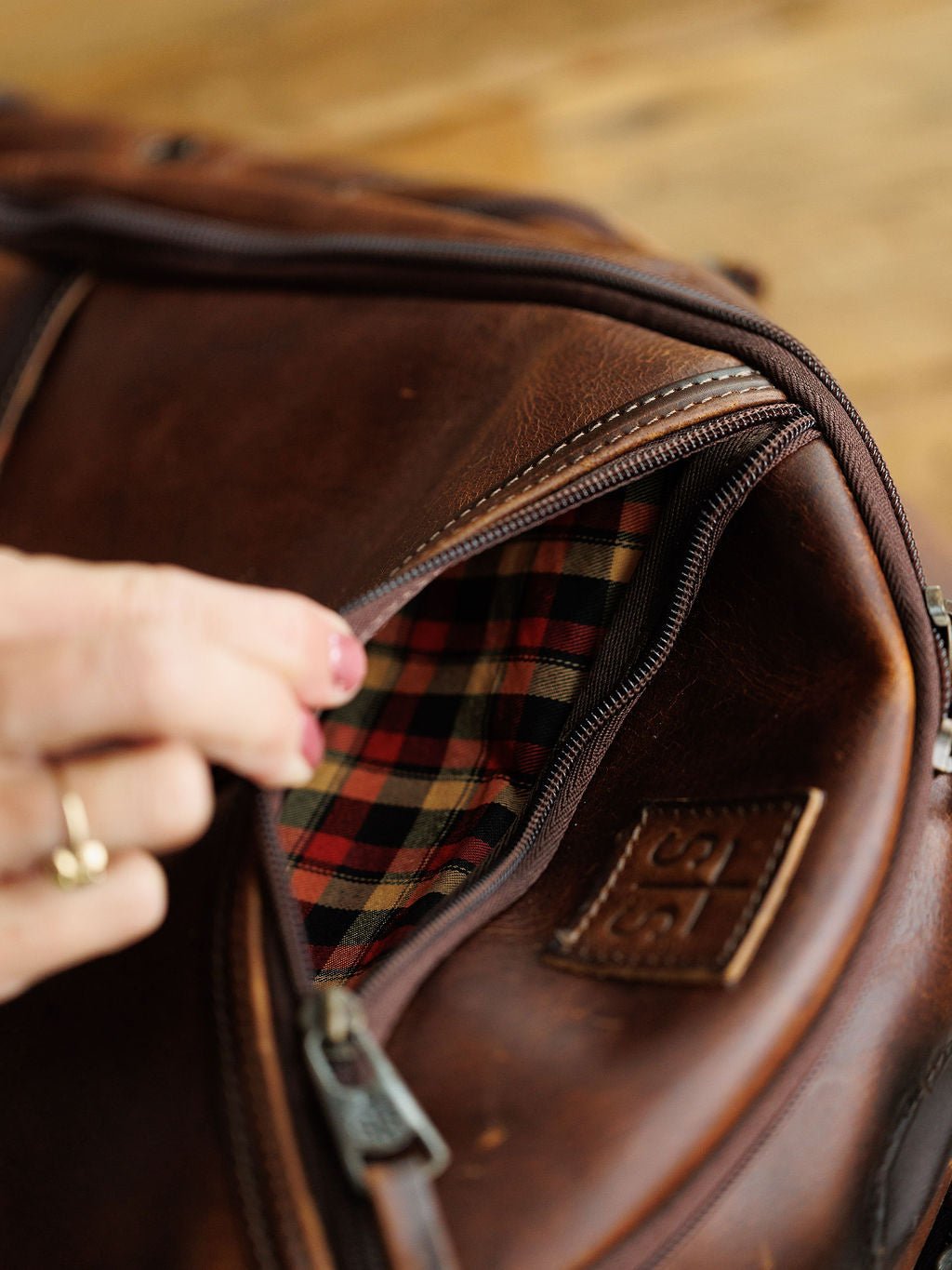 A hand with a gold ring unzips the Savannah Sevens western life{&}style Waynesboro Leather Backpack, revealing its red and black plaid fabric lining inside.