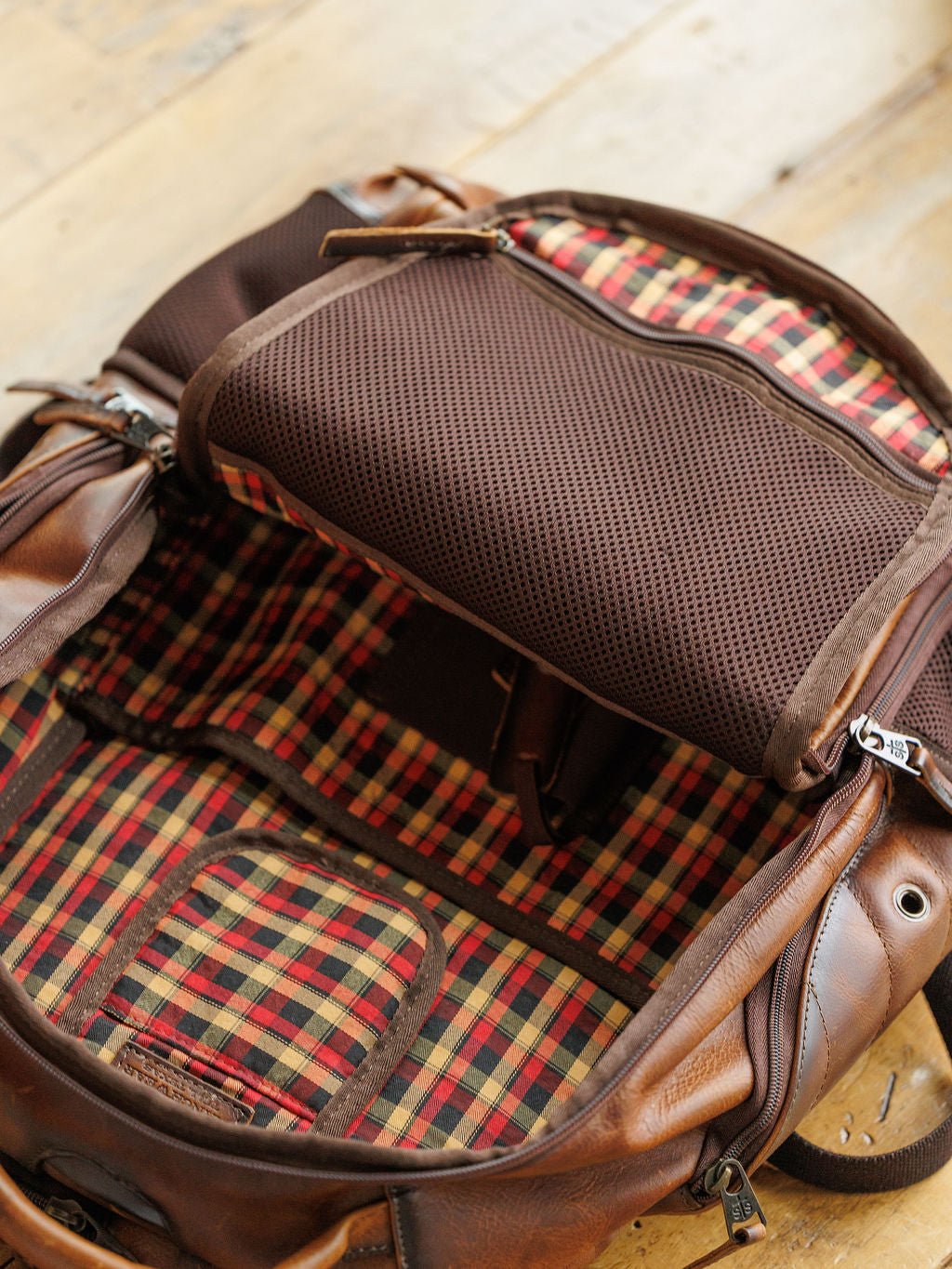 The open Waynesboro Leather Backpack by Savannah Sevens western life{&}style displays red, yellow, and black checkered lining, multiple compartments, and a brown mesh pocket on a wooden surface.