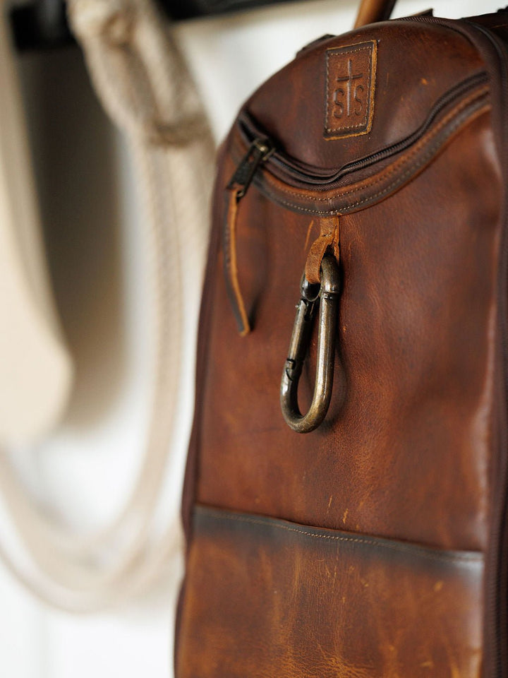 Close-up of the Waynesboro Leather Backpack by Savannah Sevens western life{&}style in brown leather, featuring a metal carabiner near the zipper, visible stitching, an embossed logo, and blurred coiled rope in the background.