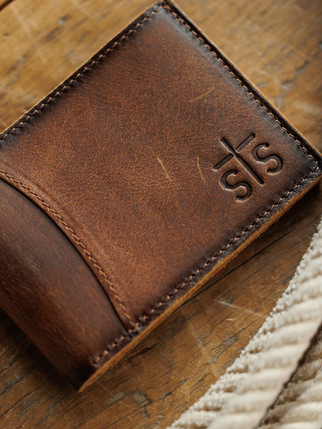 A close-up of the Waynesboro Leather Bifold Wallet by Savannah Sevens western life{&}style rests on a wooden surface, with a beige textured strap partially visible in the foreground.