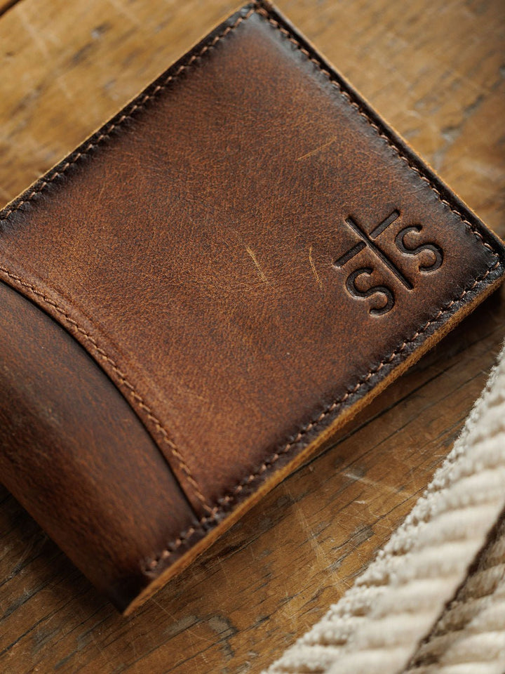 A close-up of the Waynesboro Leather Bifold Wallet by Savannah Sevens western life{&}style rests on a wooden surface, with a beige textured strap partially visible in the foreground.