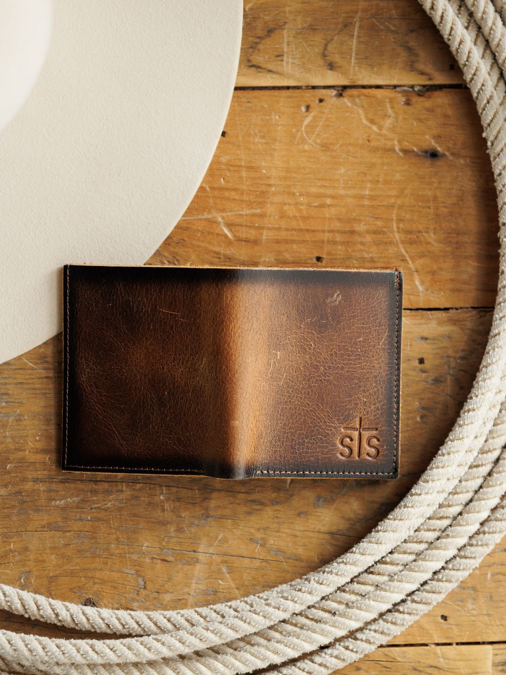 The Waynesboro Leather Hidden Cash Wallet by Savannah Sevens western life{&}style, crafted from full grain leather and embossed with "sls," sits on a rustic wooden surface beside a white cowboy hat and coiled rope.
