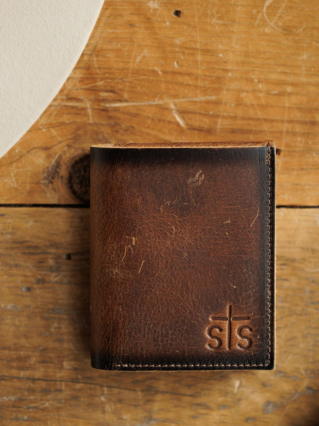 The Waynesboro Leather Hidden Cash Wallet by Savannah Sevens western life{&}style, made from worn brown full grain leather with "sts" embossed on the front, is displayed on a rustic wooden surface.