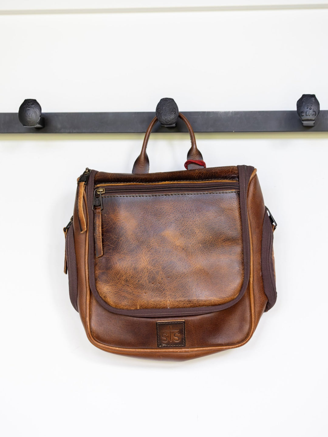 A rugged brown full grain leather Waynesboro Shave Kit by Savannah Sevens western life{&}style with a front zipper pocket hangs on a black metal wall hook rack with three hooks.